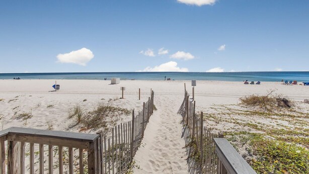 Une plage de sable avec une promenade en bois menant à la mer, un ciel dégagé et quelques personnes visibles au loin.