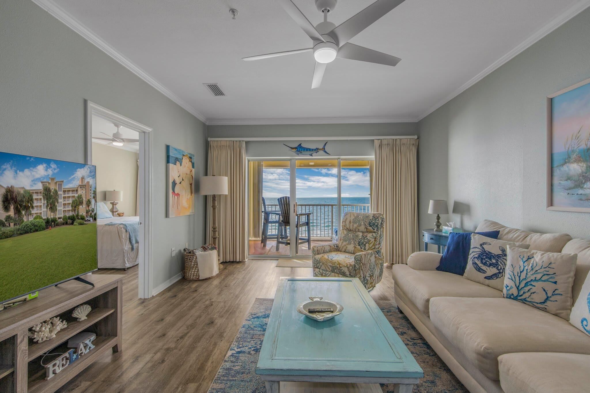 A living room with a sofa, coffee table, and TV. There is a balcony with a view of the ocean.