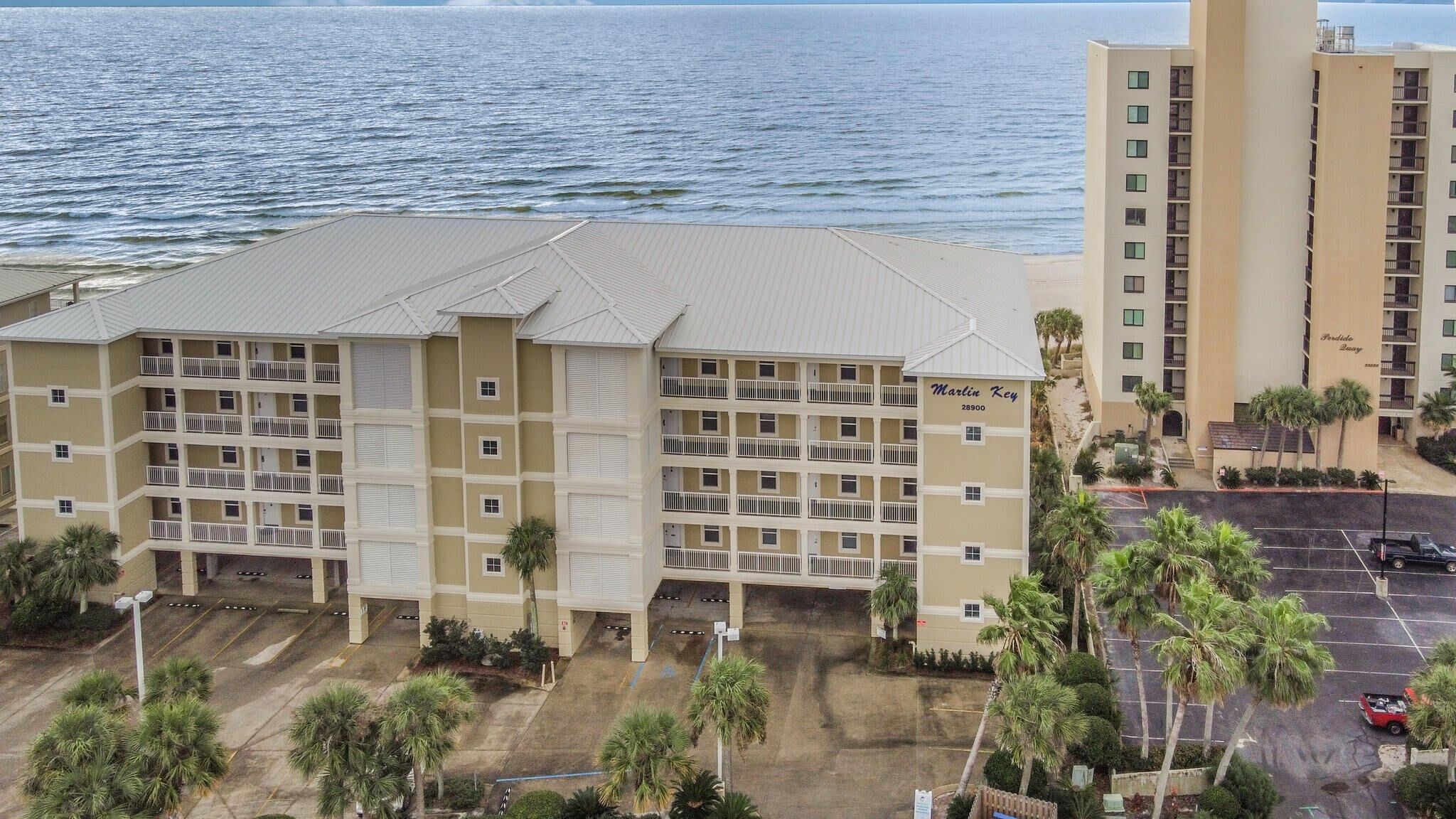 A multi-story hotel with a flat roof, multiple balconies, and a parking area with palm trees.