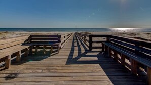 Une promenade en bois qui mène à une plage, avec des bancs et une vue dégagée sur l’océan.