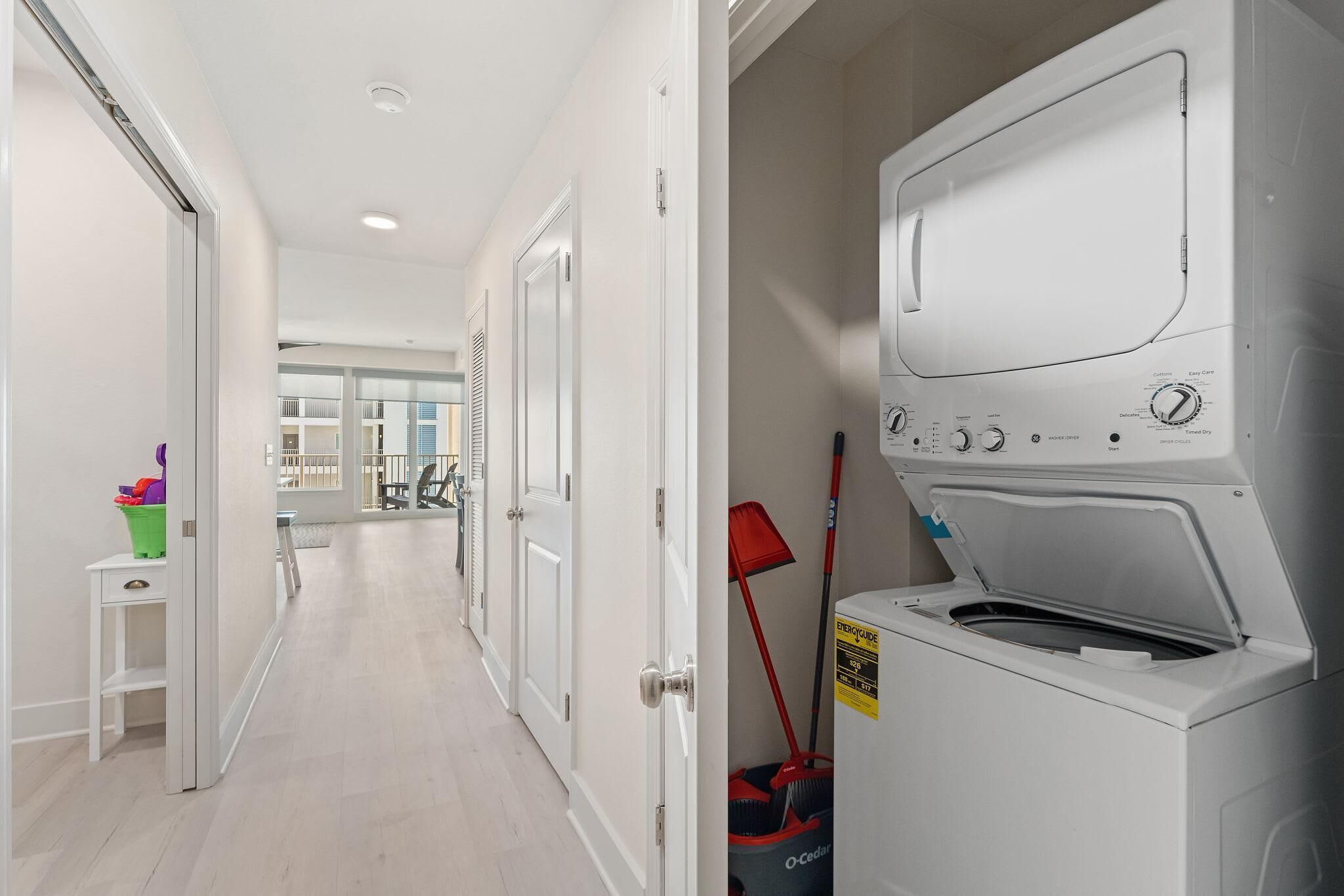 A laundry room with a front-loading washer and dryer, a green trash bin, and a red broom.
