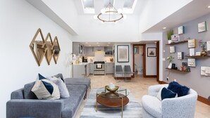 Un salon moderne avec un canapé gris, un fauteuil blanc et une table basse en verre. On aperçoit une cuisine à l’arrière-plan, avec des meubles gris et un plan de travail blanc.
