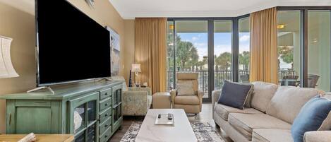 Una sala de estar con televisor de pantalla plana, un mueble verde, un sofá color beige y un balcón con vistas a palmeras.
