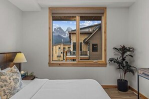A bedroom with a bed, a window with a view of buildings and mountains, a bedside table with a lamp, and a potted plant.