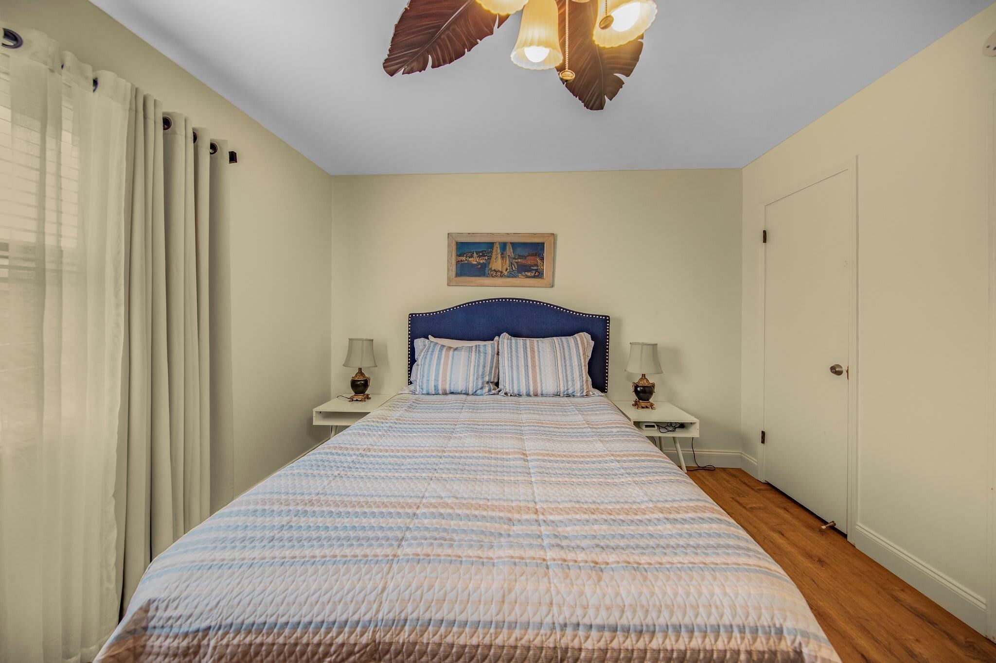 A bedroom with a bed, two bedside tables, a ceiling light fixture, a door, and a wall-mounted picture.