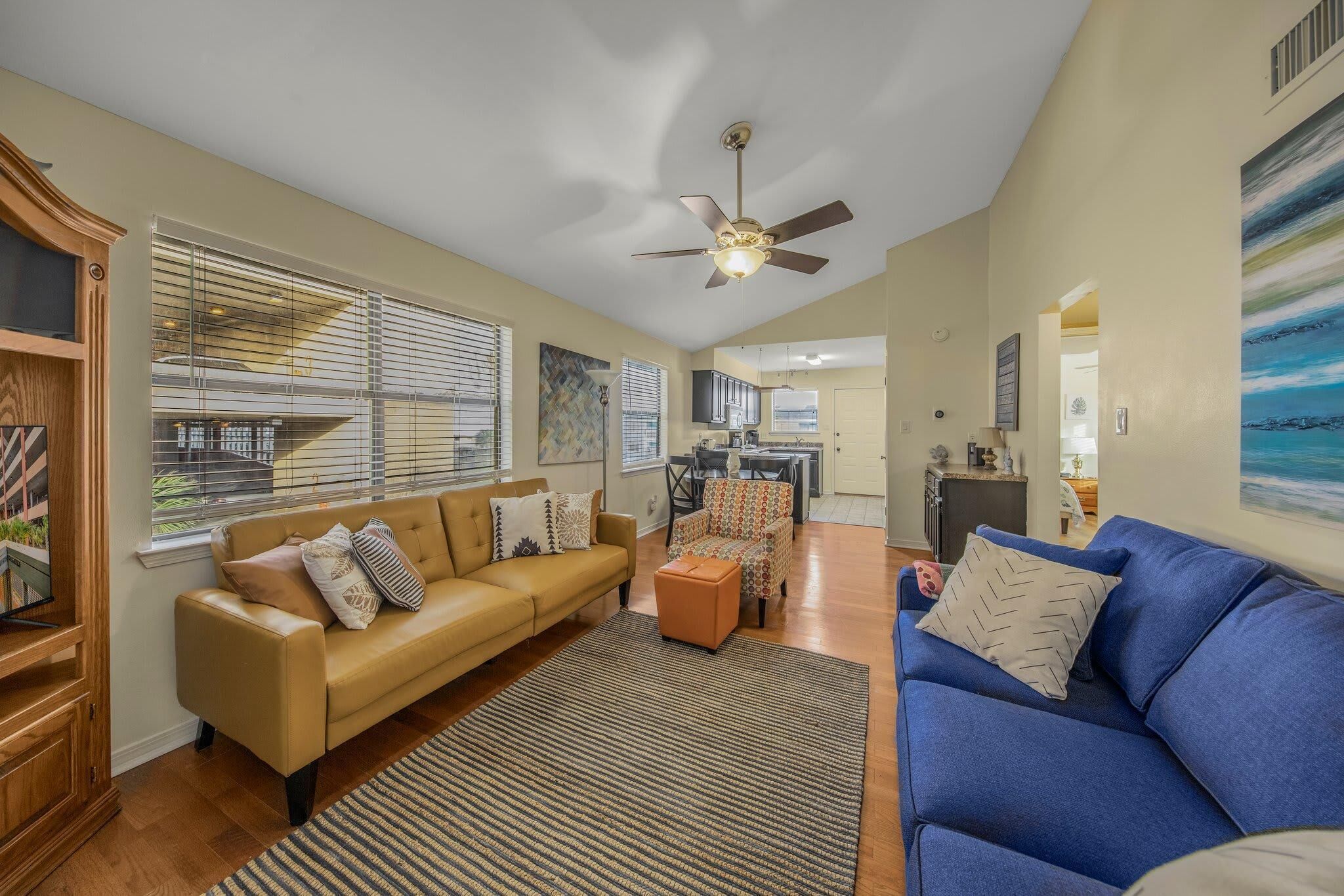 A spacious living room with a beige sofa, a blue sofa, and a patterned armchair. There is a ceiling fan, a large window with blinds, and a kitchen area in the background.