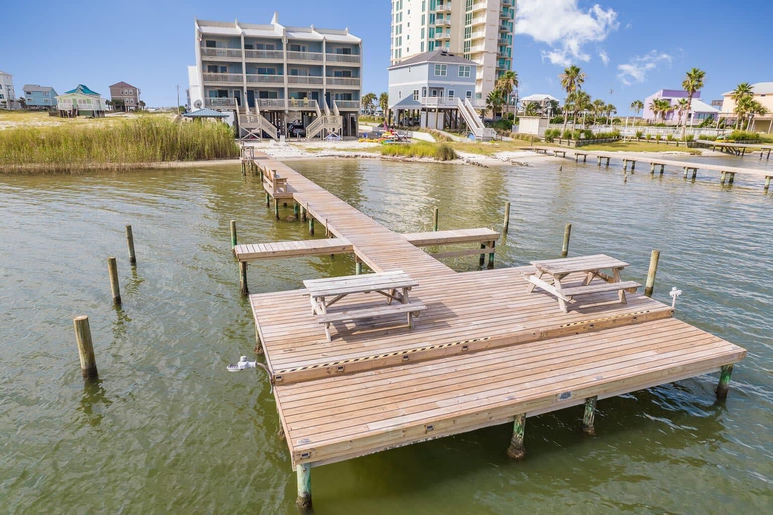 A wooden pier with picnic tables extending into a body of water, surrounded by buildings and palm trees.
