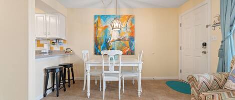 Un área de comedor con una mesa y sillas blancas, un cuadro en la pared y una cocina al fondo.