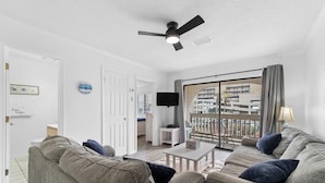Uma sala de estar com ventilador de teto, televisão e uma grande janela com vista para edifícios.