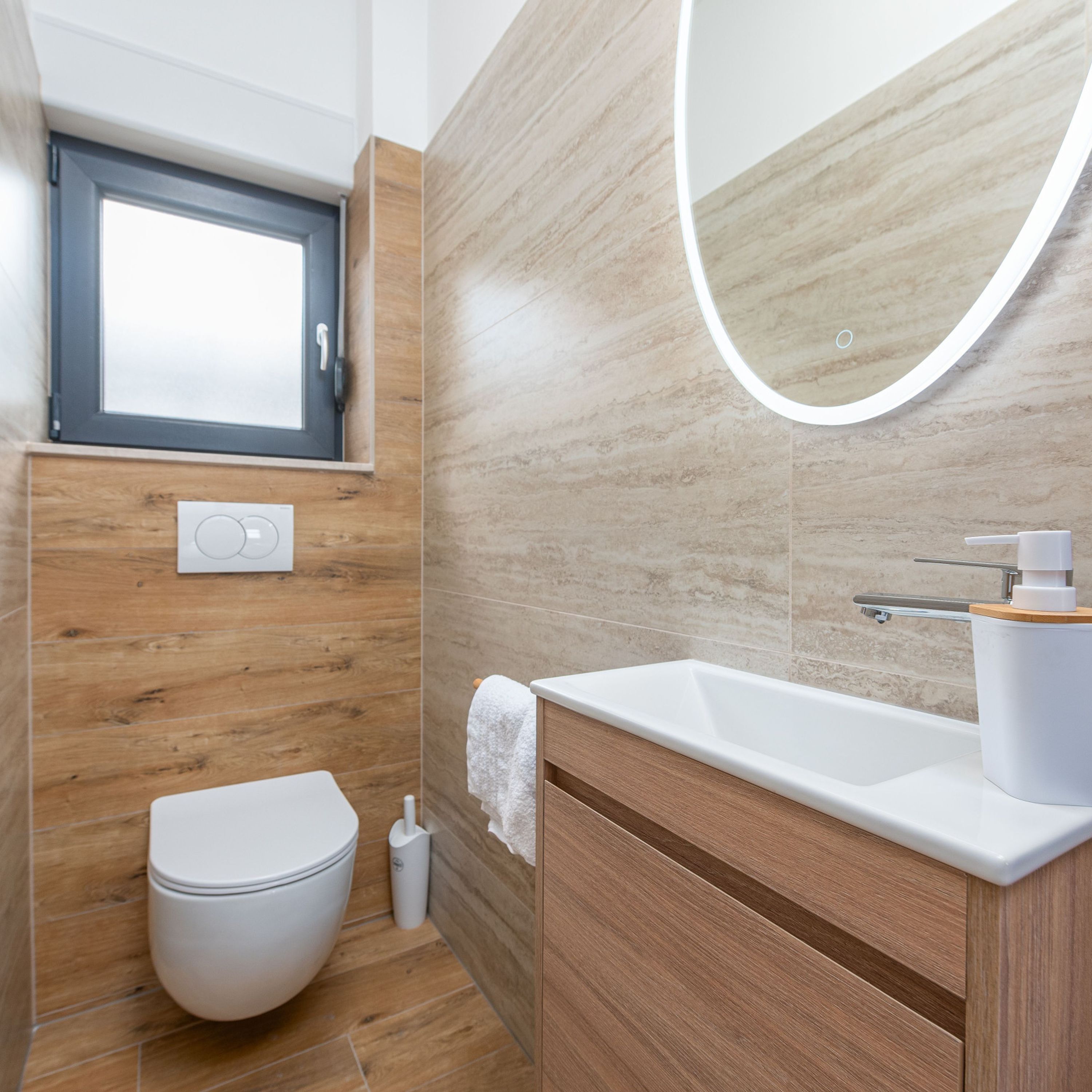 A modern bathroom with a wooden floor, a white toilet, a sink with a mirror, and a window above the toilet.