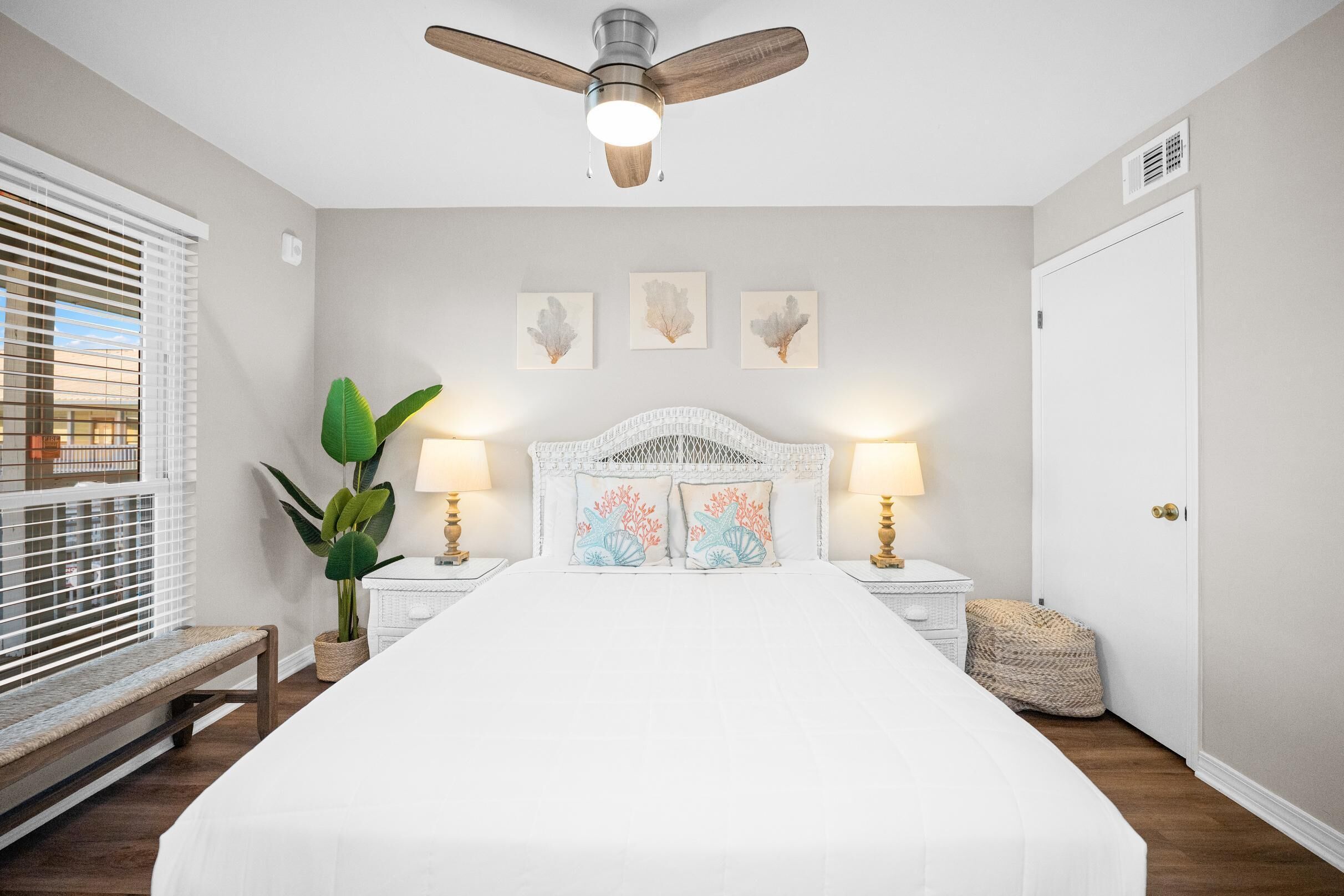 Un dormitorio con cama, ventilador de techo, lámparas de noche, decoración en la pared y una planta en maceta.