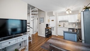 Una cocina moderna con armarios blancos, horno empotrado y refrigerador. Hay una mesa de comedor con sillas y un televisor de pantalla plana sobre un mueble blanco.