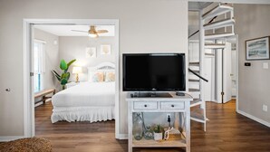Un dormitorio con cama, televisor sobre una consola blanca, escalera y una puerta que da a otra habitación.