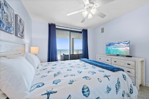 Quarto com cama, televisão, ventoinha no teto e janela com cortinas.