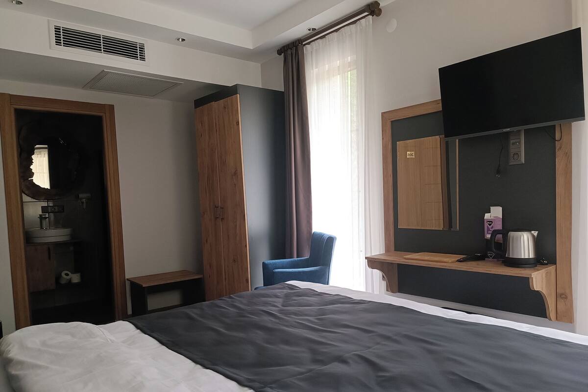 A bedroom with a bed, a TV mounted on the wall, a wardrobe, a chair, and a small table with a kettle.