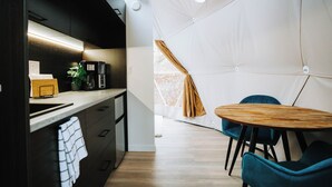 A compact kitchen with modern appliances, a dining area with a round wooden table and blue chairs, and a tent-like structure with a window and curtain. - Oak Haven Oasis ~ Dome 1 (Brandon)