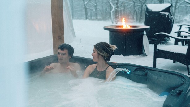 Un bain à remous où se trouvent deux personnes, entouré de neige.