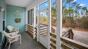 A porch with wicker chairs, a wooden railing, and a view of trees and a gravel path. - The Rookery 3504 (Gulf Shores)