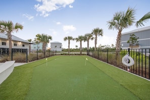 A croquet field with artificial turf, surrounded by a black metal fence, palm trees, and buildings in the background.