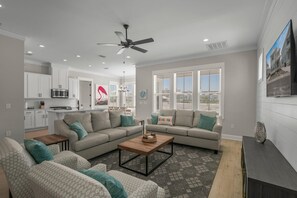 A spacious living room with a large sectional sofa, a coffee table, and a TV on a stand. There is a kitchen area with white cabinets and a microwave. - Summer Salt 24170 (Orange Beach)
