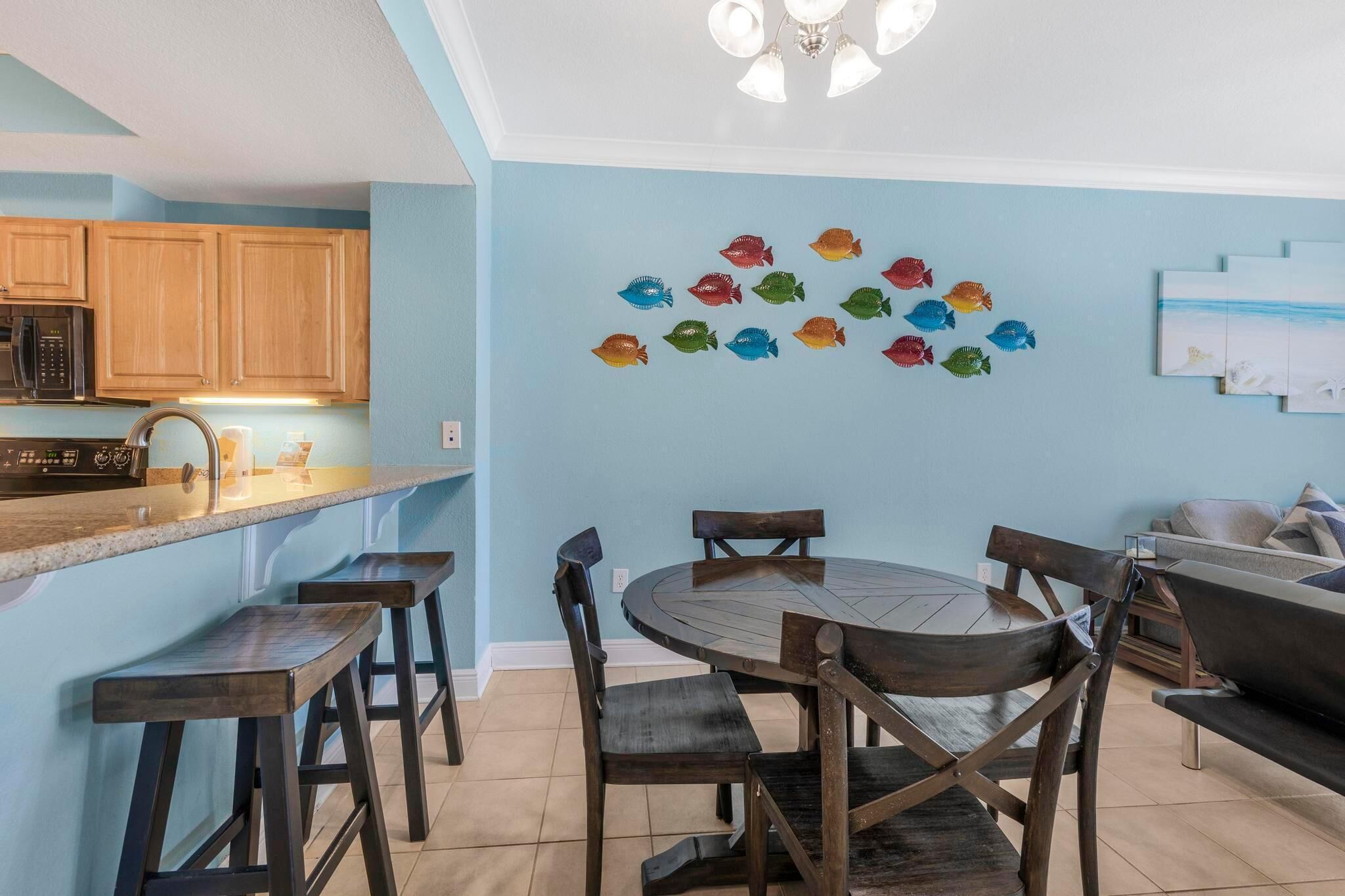 Une cuisine avec des meubles en bois, un four à micro-ondes et un coin bar avec des tabourets. On y trouve une table à manger avec des chaises et une décoration murale représentant des poissons.