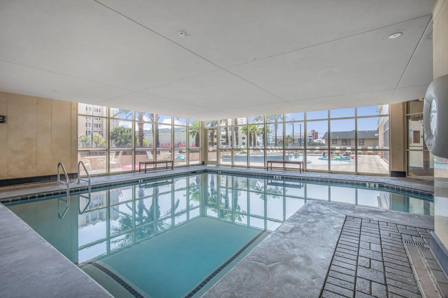 An indoor swimming pool with clear glass walls, a tiled floor, and a bench by the pool.