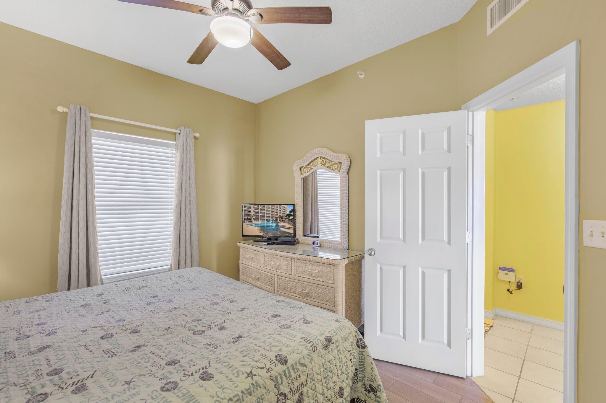 A bedroom with a bed, a dresser, a television, a ceiling fan, and a door leading to another room.