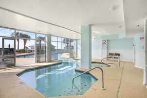 Una piscina interior con paredes de vidrio transparente, suelo de baldosas y una zona de estar junto a la piscina con sillas y un parasol.