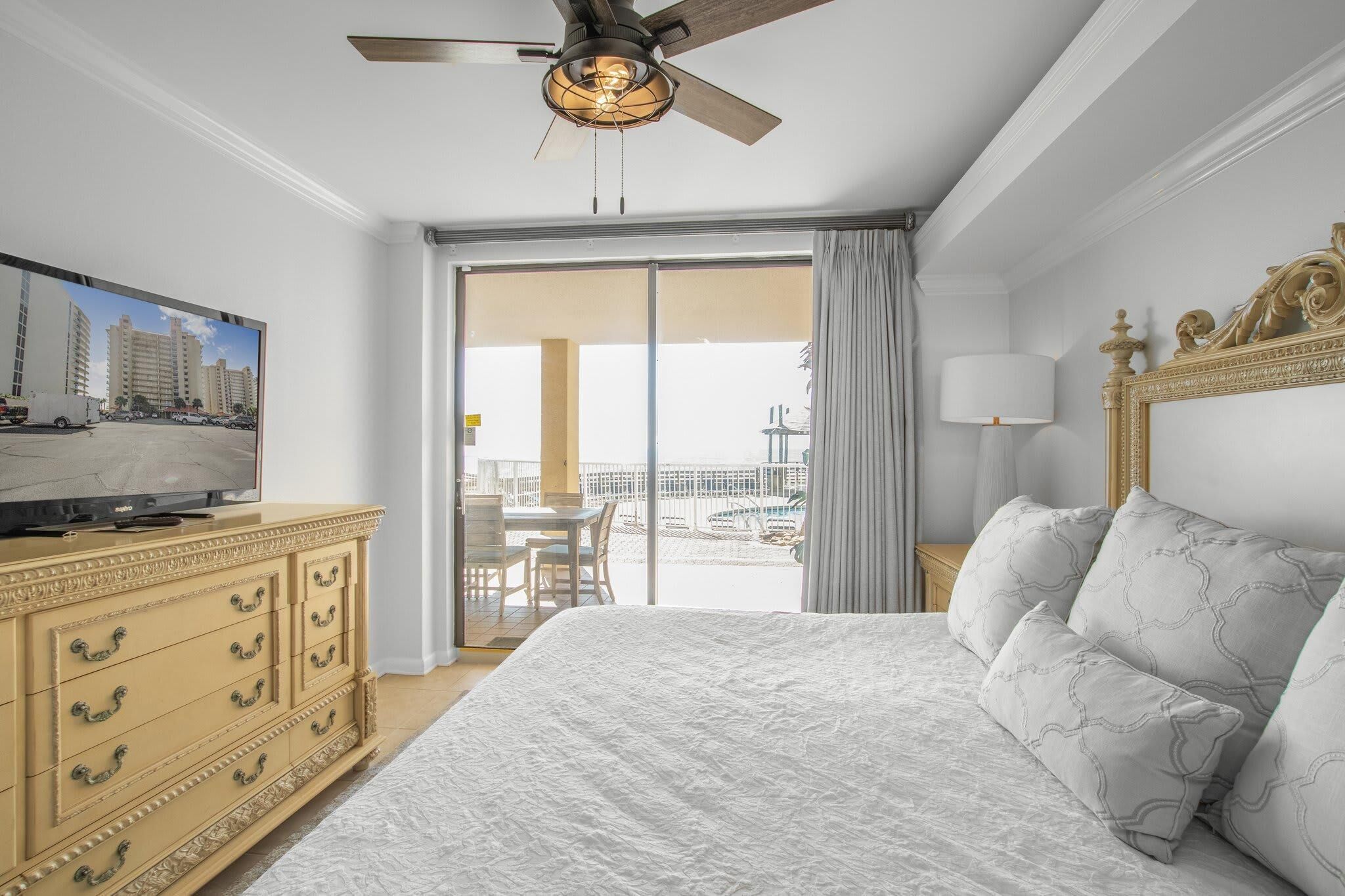 Ein Hotelzimmer mit einem großen Bett, einem Fernseher, einem Deckenventilator und einem Balkon mit Blick auf Gebäude und einen Parkplatz.
