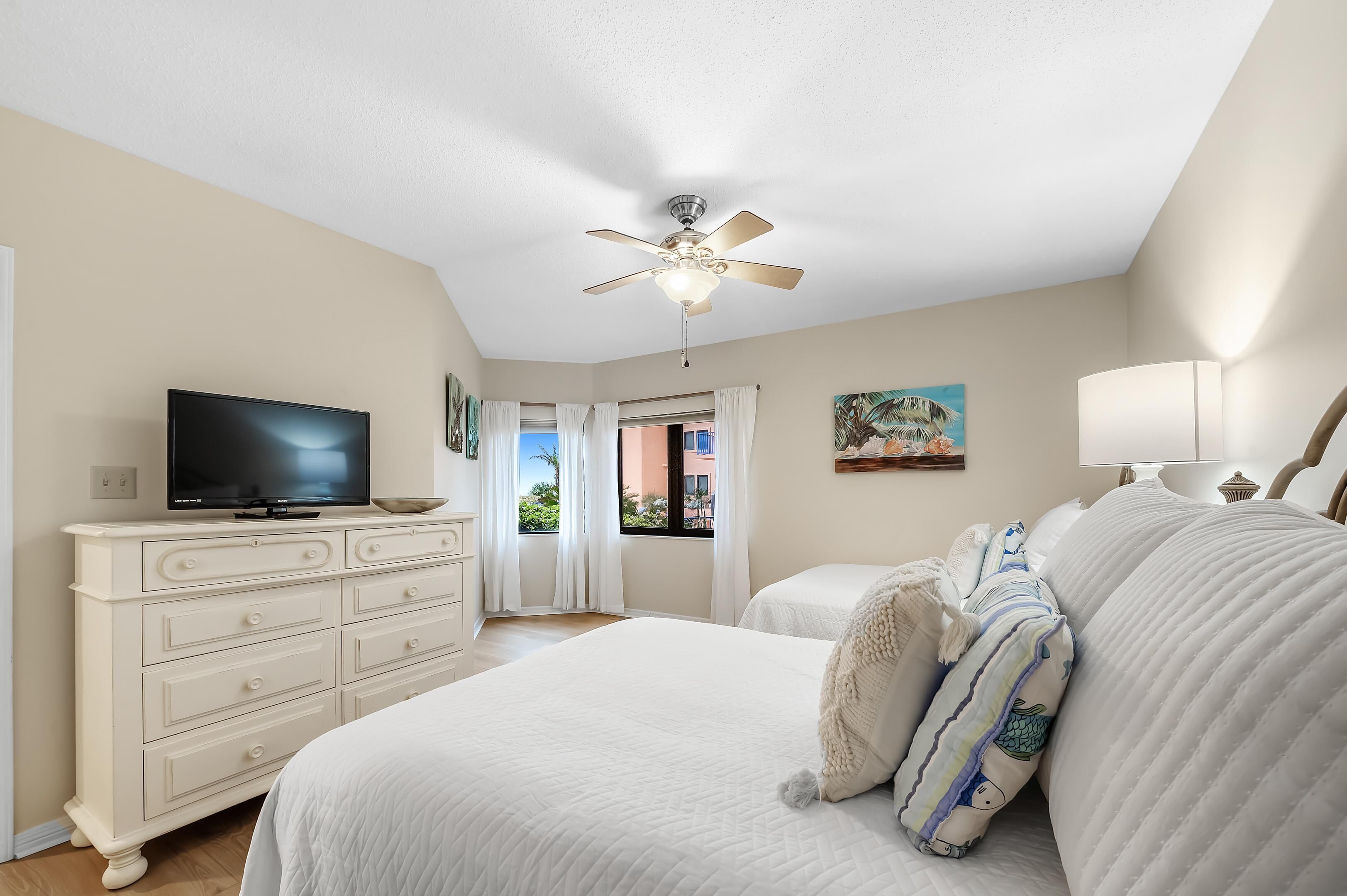 A bedroom with a bed, a dresser, a television, a ceiling fan, and a window with curtains.
