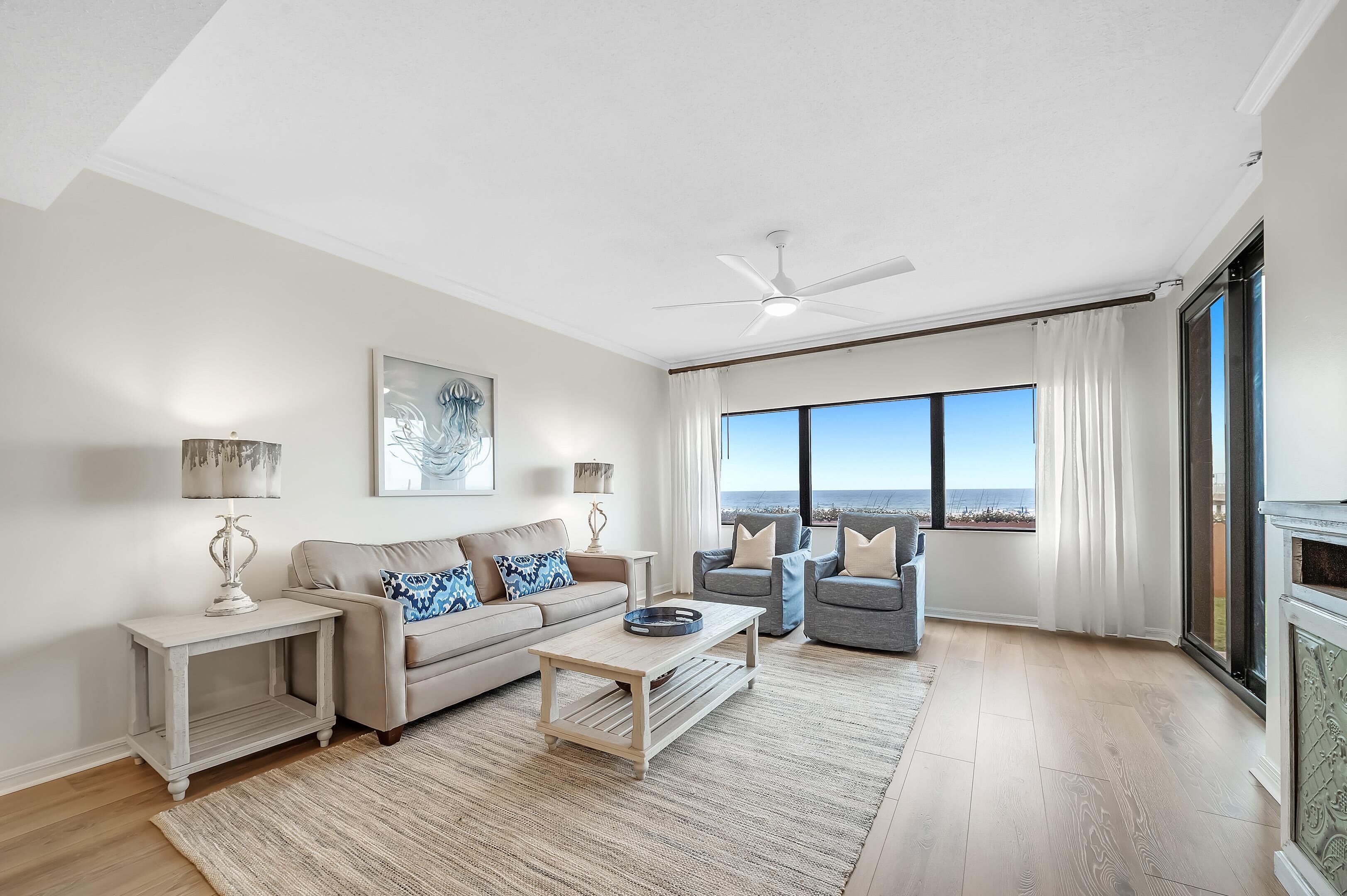 A spacious living room with a high ceiling, a ceiling fan, a large window with a view of the ocean, a beige sofa, a gray armchair, and a coffee table.