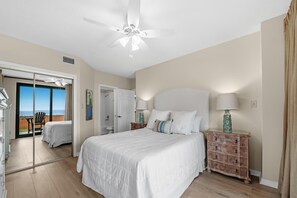 A bedroom with a large bed, a ceiling fan, a nightstand with a lamp, and a view of the ocean through a sliding door. - Four Seasons 103e (Orange Beach)