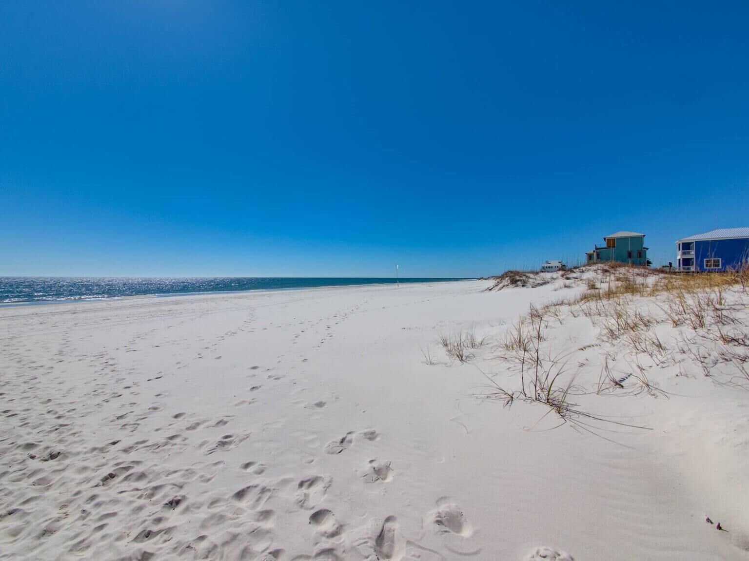 A beach with footprints in the sand, a dune, and a building with a blue roof.