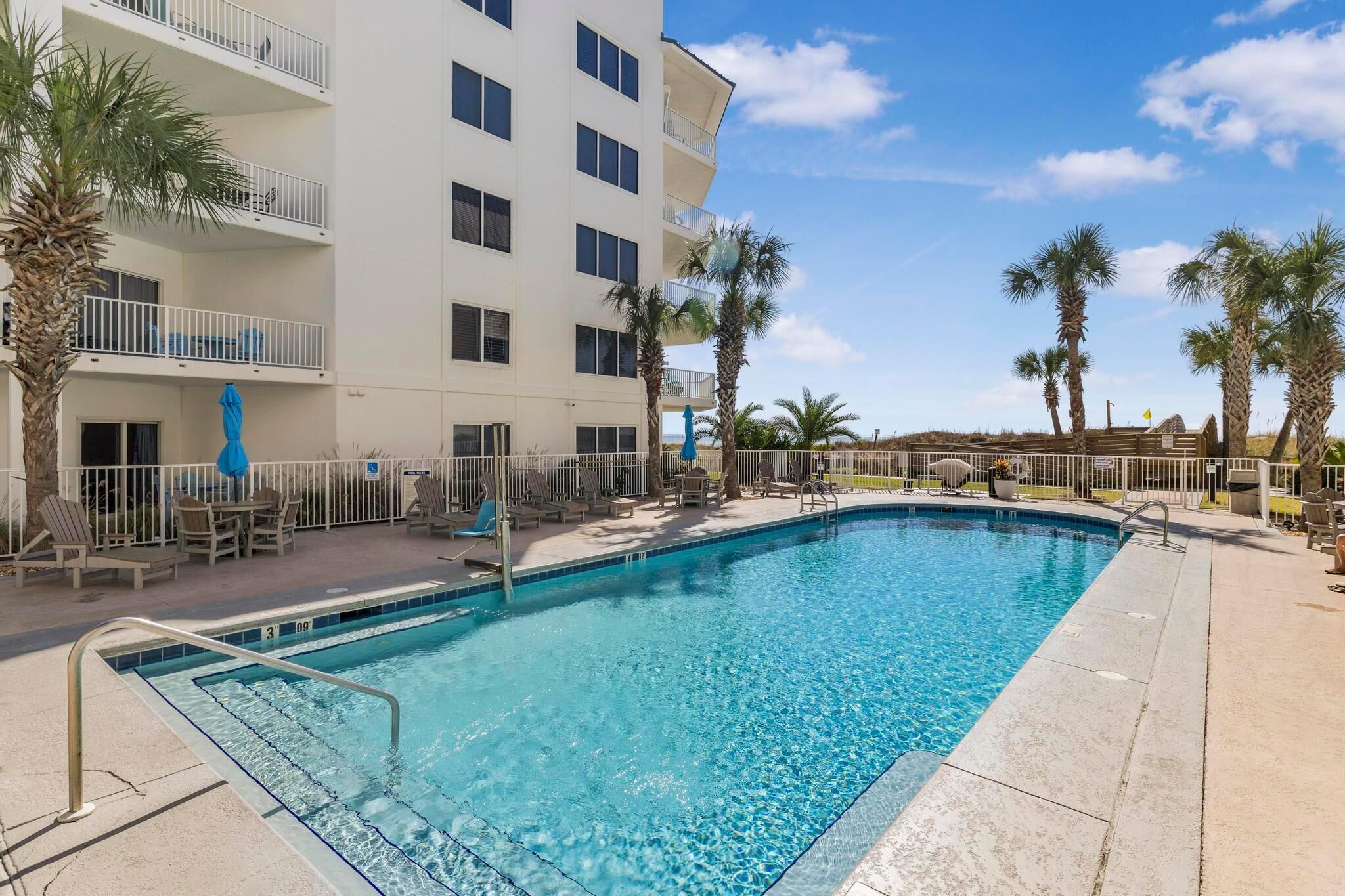 A swimming pool with lounge chairs and umbrellas, surrounded by a white building with balconies and palm trees.