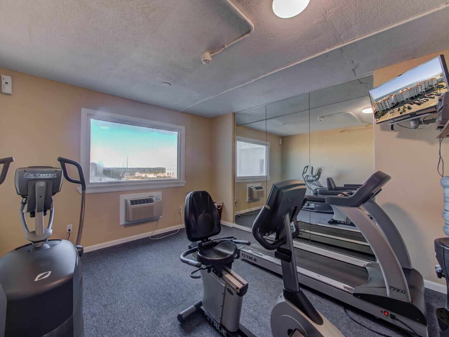 Une salle de sport équipée de tapis de course, d’un vélo elliptique et d’un vélo d’appartement. Des fenêtres offrent une vue sur les bâtiments et un grand miroir est fixé au mur.