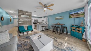 Una sala moderna con una mesa de centro blanca, sillas color aguamarina y una zona de cocina con armarios blancos y un taburete de bar.
