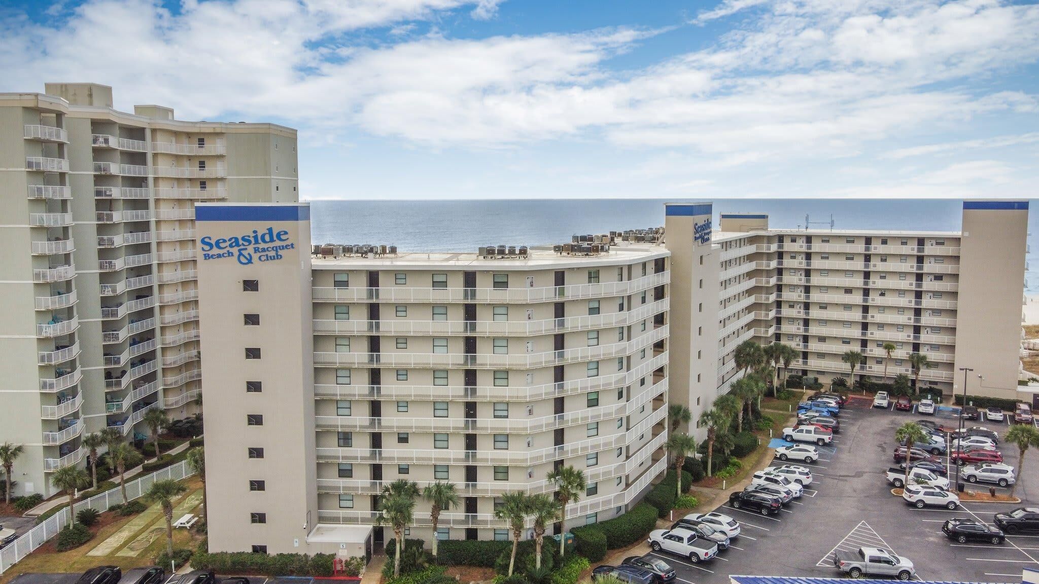 A multi-story hotel with a beach and racquet club sign, parking lot, and ocean view.