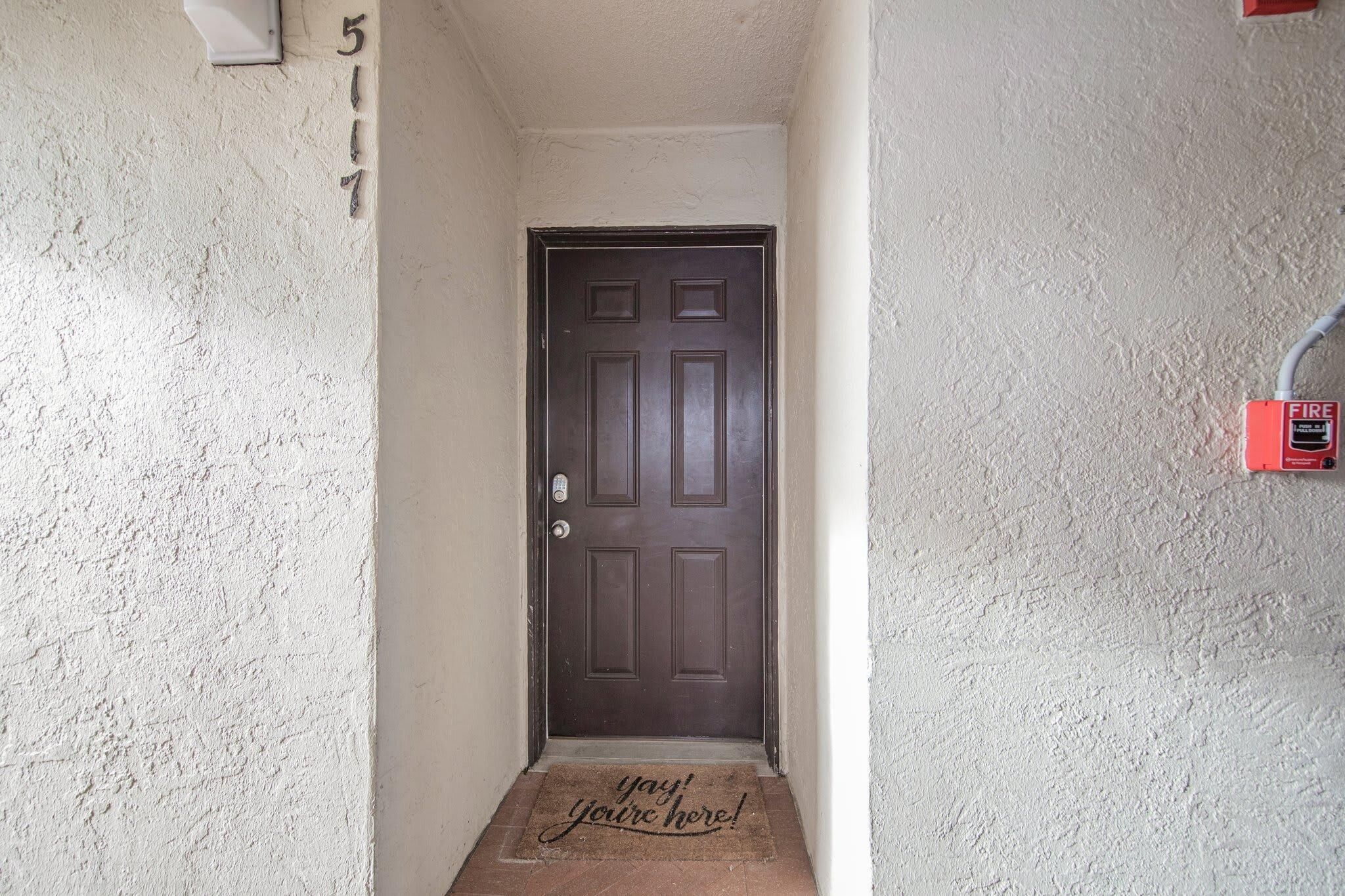 A narrow hallway with a brown door, a doormat with the text "yay! you're here!", and a fire alarm on the right wall.