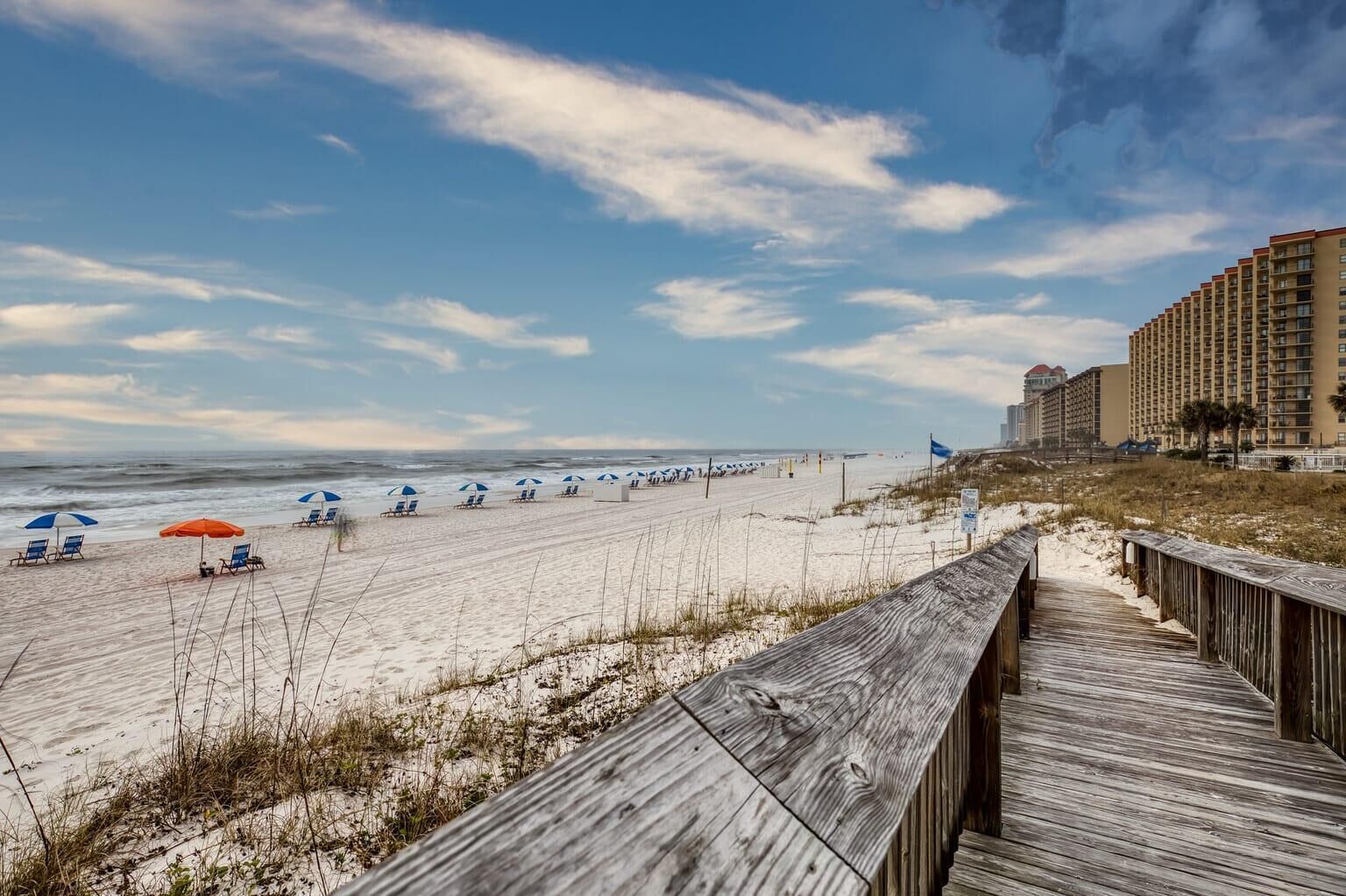 A beach with rows of beach chairs and umbrellas, a wooden boardwalk, and a hotel in the background.