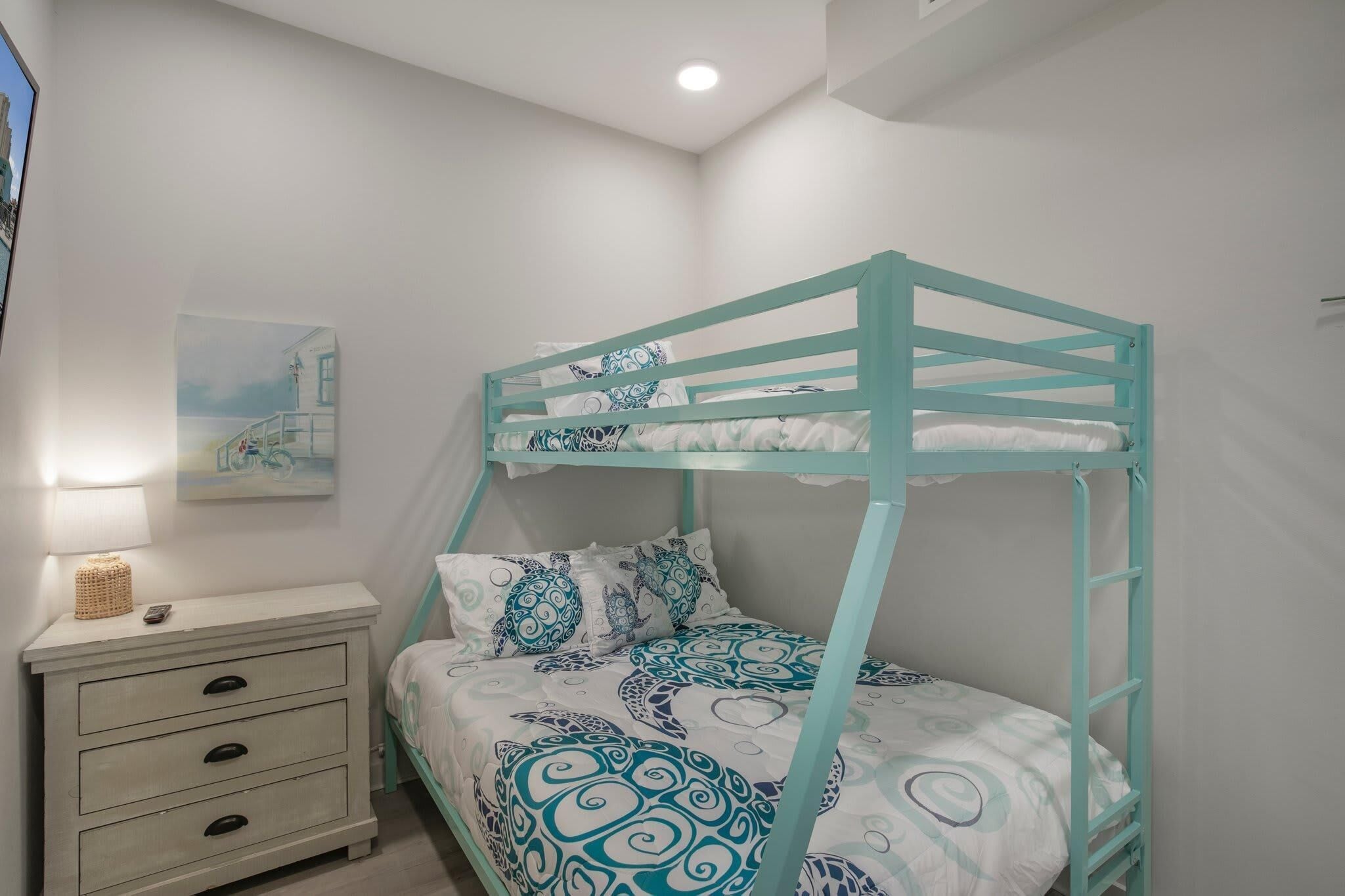 A bunk bed with a teal frame and patterned bedding, a bedside table with a lamp, and a wall-mounted painting.
