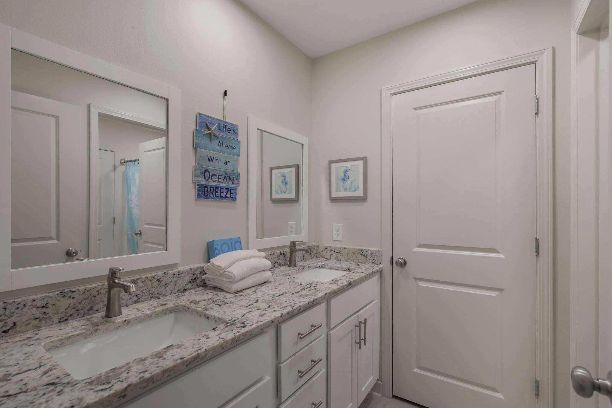 A bathroom with two sinks, a large mirror, and a door. There are framed pictures on the wall and a sign with text.