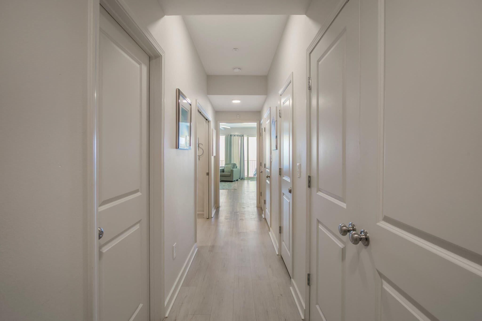 A long, narrow hallway with white doors on both sides, leading to a bright room at the end.