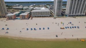 Ein Strand mit Reihen blauer Sonnenschirme und einigen Personen. Im Hintergrund sind mehrstöckige Gebäude und ein Gewässer zu sehen.