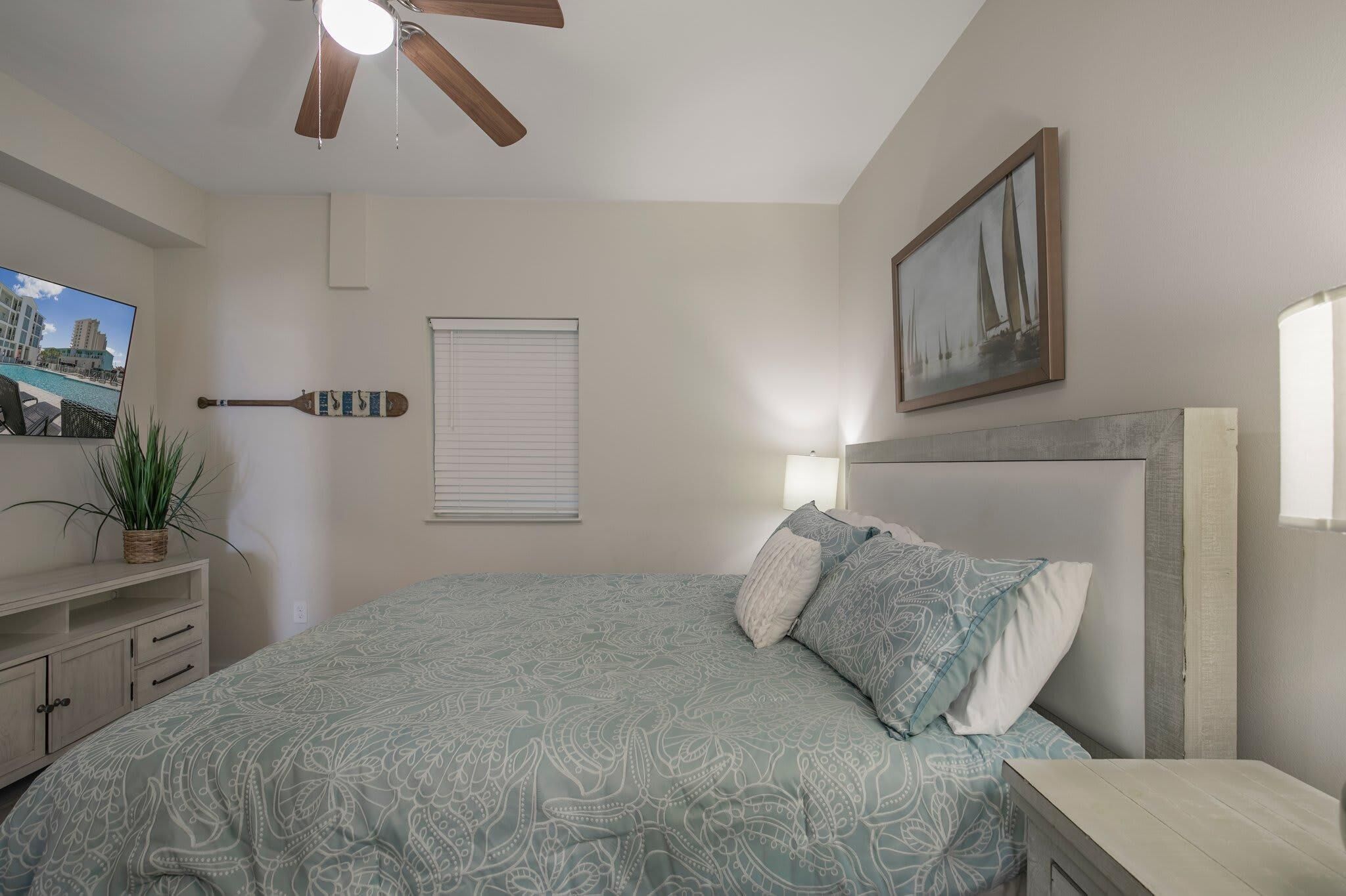 Une chambre à coucher avec un lit, un ventilateur de plafond, une télévision, une plante, une fenêtre avec des stores et un tableau au mur.
