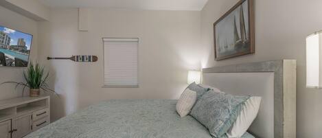 A bedroom with a bed, a ceiling fan, a TV, a plant, a window with blinds, and a painting on the wall.