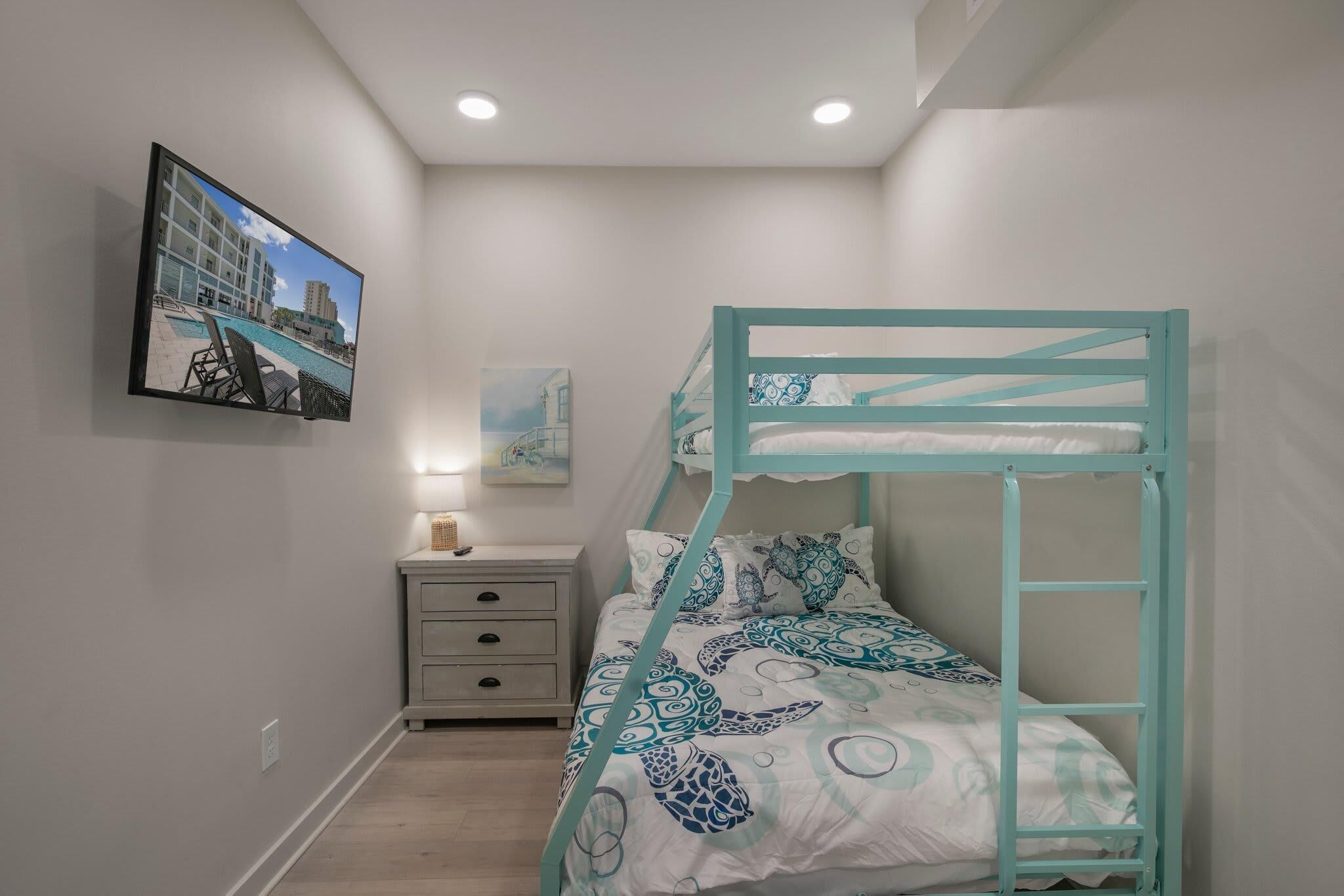 Un lit superposé avec une structure de couleur turquoise et une literie à motifs, une table de chevet avec une lampe, une petite commode et un tableau représentant un bâtiment.