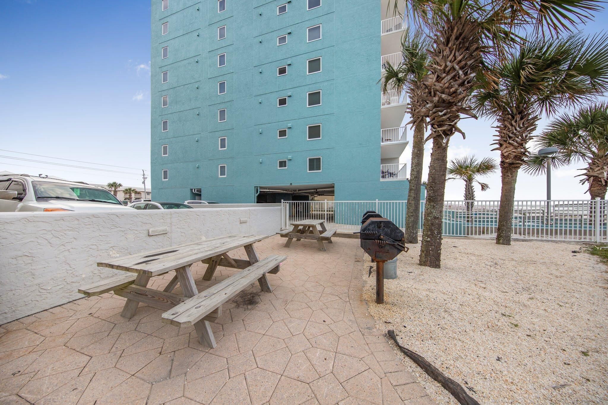 A beachside patio with picnic tables, a barbecue grill, and a tall blue building in the background.