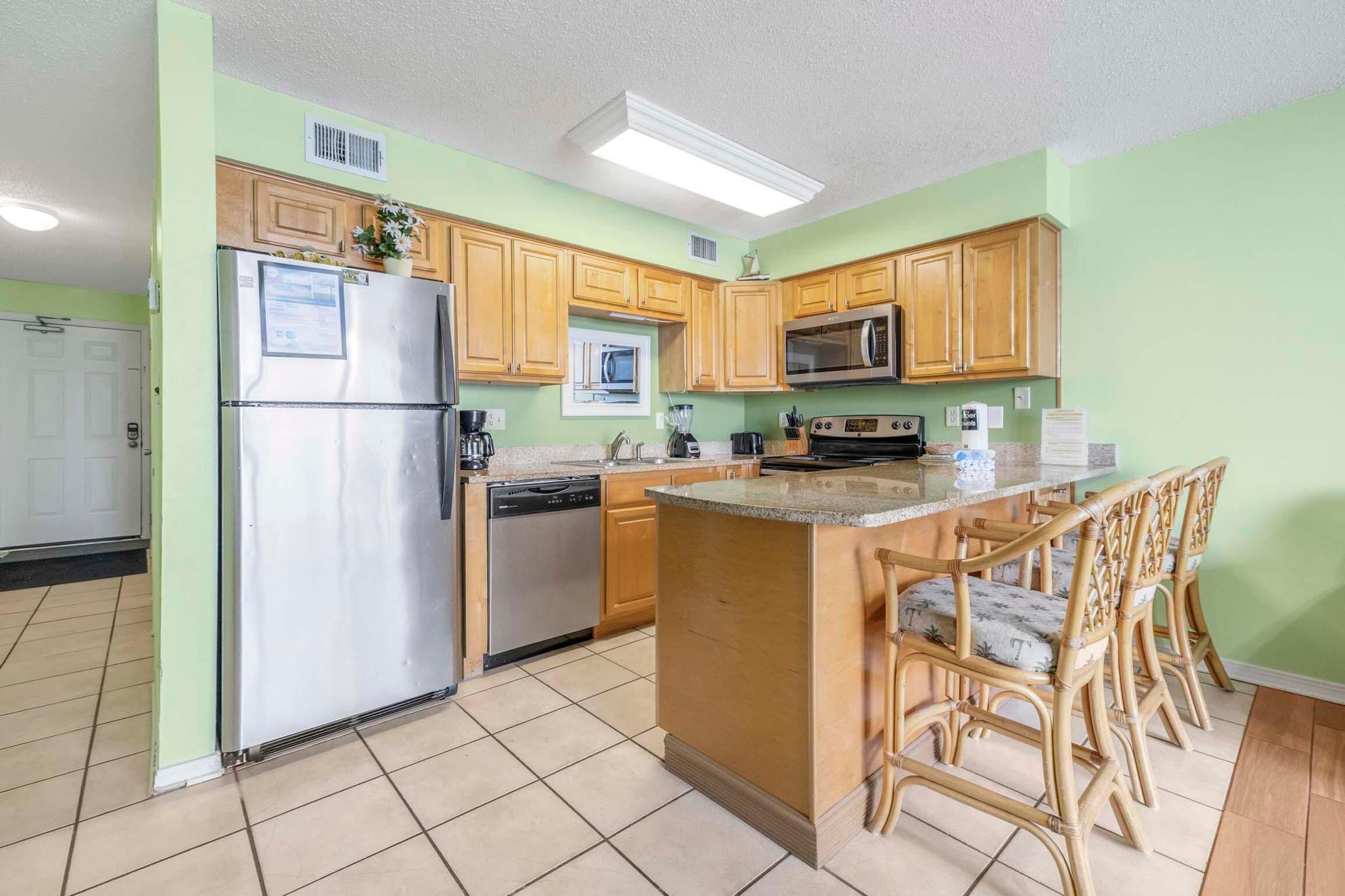 A kitchen with wooden cabinets, a refrigerator, a microwave, a dishwasher, and a bar area with chairs.
