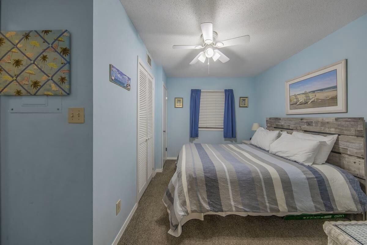 A bedroom with a bed, a ceiling fan, a window with blinds, and a beach-themed artwork on the wall.