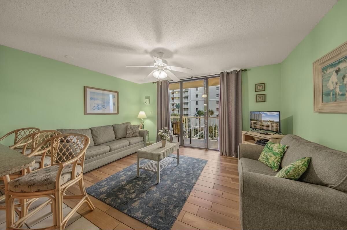 A living room with a sofa, a coffee table, and a dining table with chairs. There is a ceiling fan, a television, and a window with curtains.